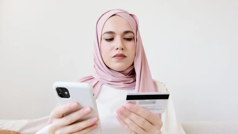 A woman in a hijab uses her smartphone and credit card for online shopping while sitting indoors.