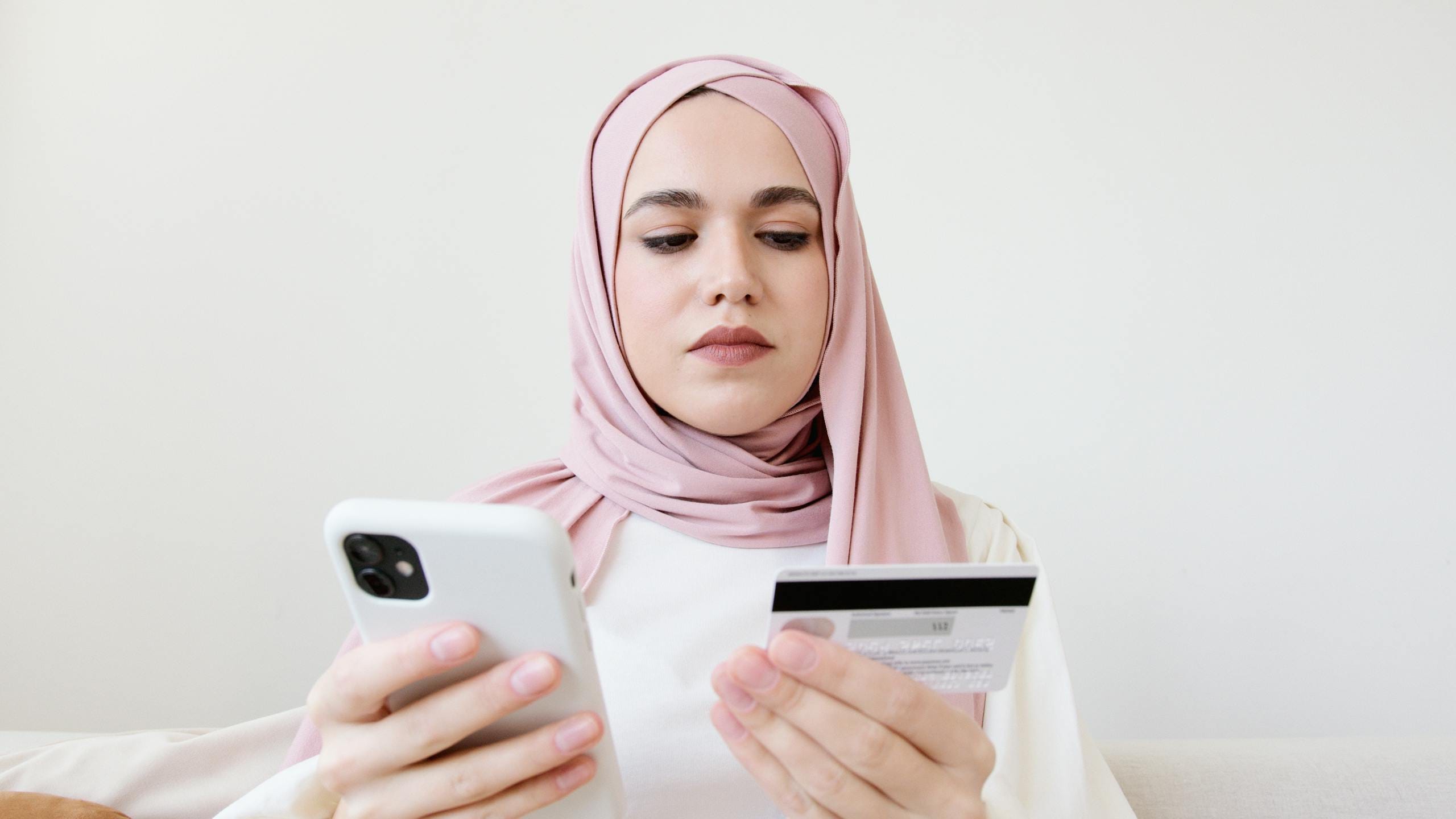A woman in a hijab uses her smartphone and credit card for online shopping while sitting indoors.